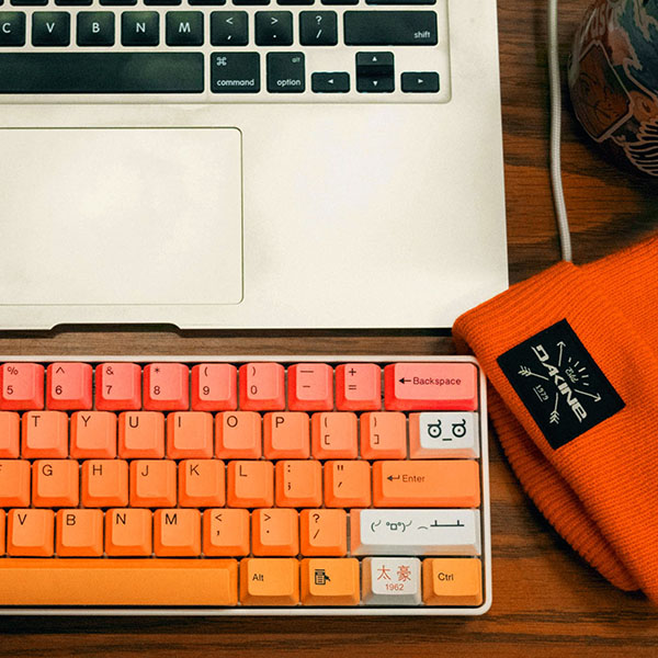 An overhead shot of a laptop and keyboard with pink-orange-yellow keys, alongside an orange beanie